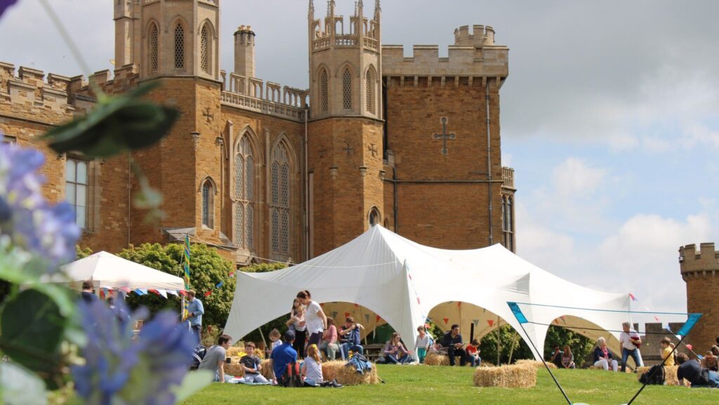 People sit and gather on a grassy lawn around a white open-sided marquee, with hay bales used as seating. A large historic brick building with towers and arched windows stands in the background, while flowers and greenery appear in the foreground under a bright sky.