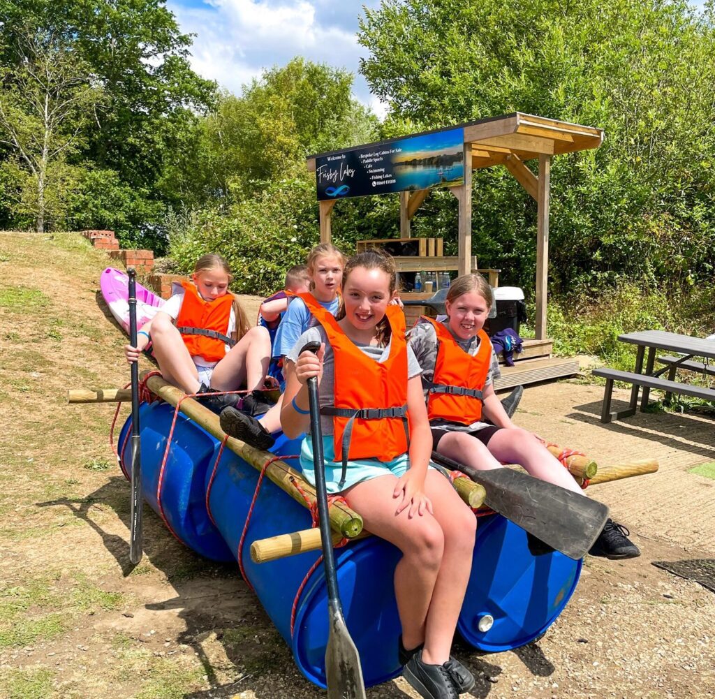 Participants wearing life jackets sit on a homemade barrel raft holding paddles at Frisby Lakes.