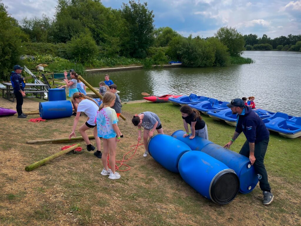 Children and instructors build a barrel raft beside a lakeside activity area.