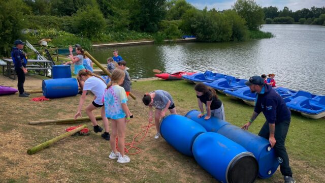 Children and instructors build a barrel raft beside a lakeside activity area.