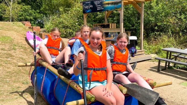 Participants wearing life jackets sit on a homemade barrel raft holding paddles at Frisby Lakes.