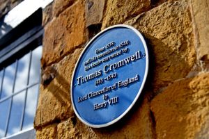 A blue circular heritage plaque mounted on a weathered stone wall, stating that Thomas Cromwell lived there between 1530 and his execution in 1540; a window with black framing is partially visible to the left.