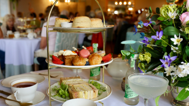 Three-tier afternoon tea stand on a white tablecloth with finger sandwiches, scones, pastries, strawberries, and a pink macaron, alongside cups of tea and a pale cocktail. A floral arrangement sits to the right, with a softly blurred dining room and seated guests in the background.