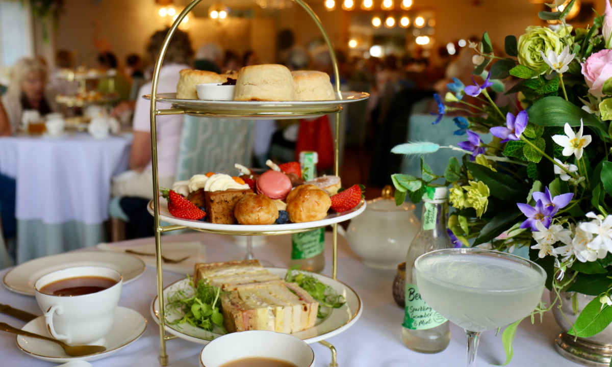 Three-tier afternoon tea stand on a white tablecloth with finger sandwiches, scones, pastries, strawberries, and a pink macaron, alongside cups of tea and a pale cocktail. A floral arrangement sits to the right, with a softly blurred dining room and seated guests in the background.