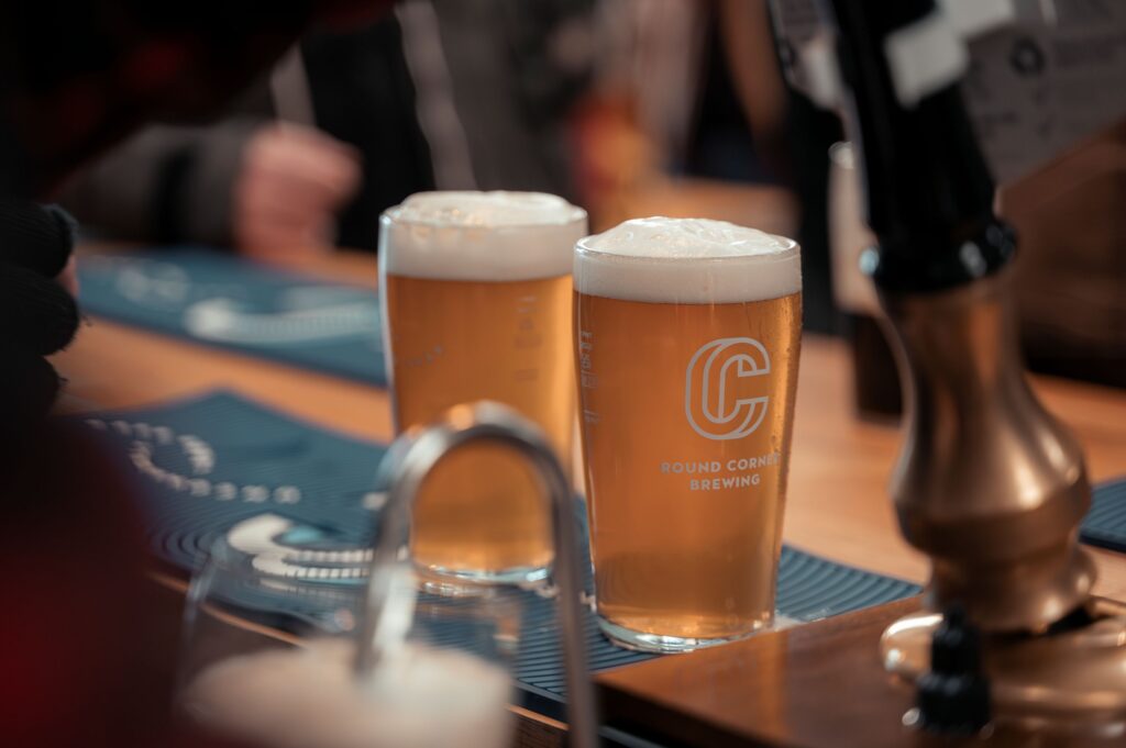 Two pints of pale beer with foamy heads sit on a wooden bar inside Round Corner Brewing’s taproom. The branded glasses are in focus, while bar fixtures and patrons in the background are softly blurred.