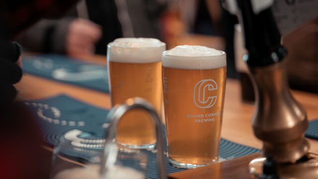 Two pints of pale beer with foamy heads sit on a wooden bar inside Round Corner Brewing’s taproom. The branded glasses are in focus, while bar fixtures and patrons in the background are softly blurred.