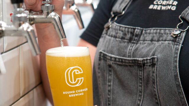 Close-up of a brewery staff member pouring a pale golden beer from a tap into a branded Round Corner Brewing pint glass. Multiple beer taps line a white tiled wall in the background, and the beer has a thick white head.