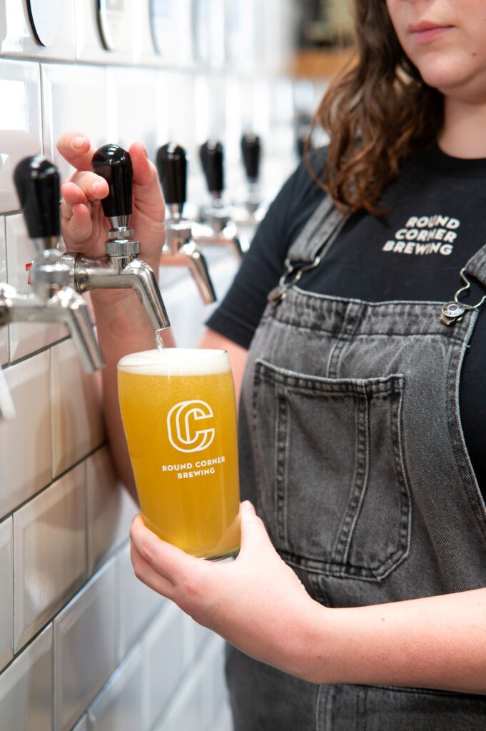 Close-up of a brewery staff member pouring a pale golden beer from a tap into a branded Round Corner Brewing pint glass. Multiple beer taps line a white tiled wall in the background, and the beer has a thick white head.