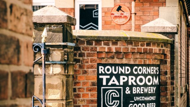 Entrance to Round Corner’s Taproom & Brewery, viewed through a narrow brick-lined passageway. A black painted sign on a curved red-brick wall reads “Round Corner’s Taproom & Brewery” with an arrow pointing toward the entrance. The brewery building and a tiled roof are visible behind the wall in bright daylight.