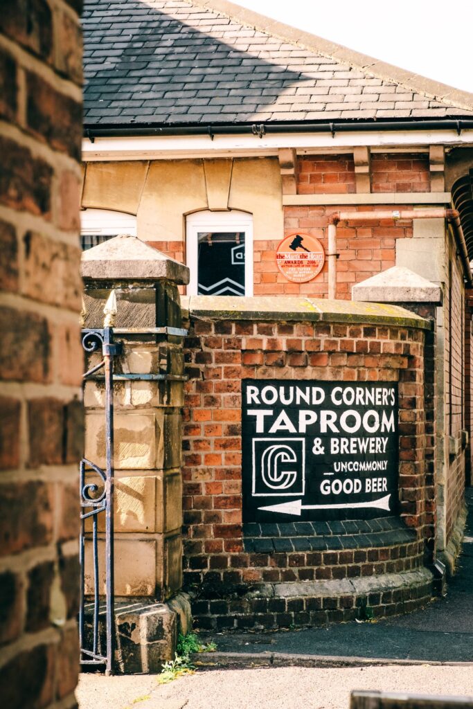 Entrance to Round Corner’s Taproom & Brewery, viewed through a narrow brick-lined passageway. A black painted sign on a curved red-brick wall reads “Round Corner’s Taproom & Brewery” with an arrow pointing toward the entrance. The brewery building and a tiled roof are visible behind the wall in bright daylight.