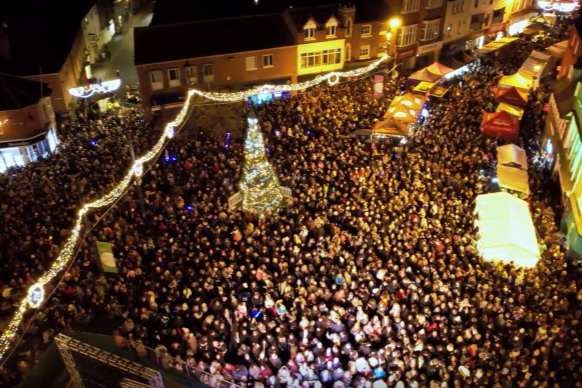 Aerial view of a large crowd gathered around a brightly lit Christmas tree and festive market stalls in a town centre at night.