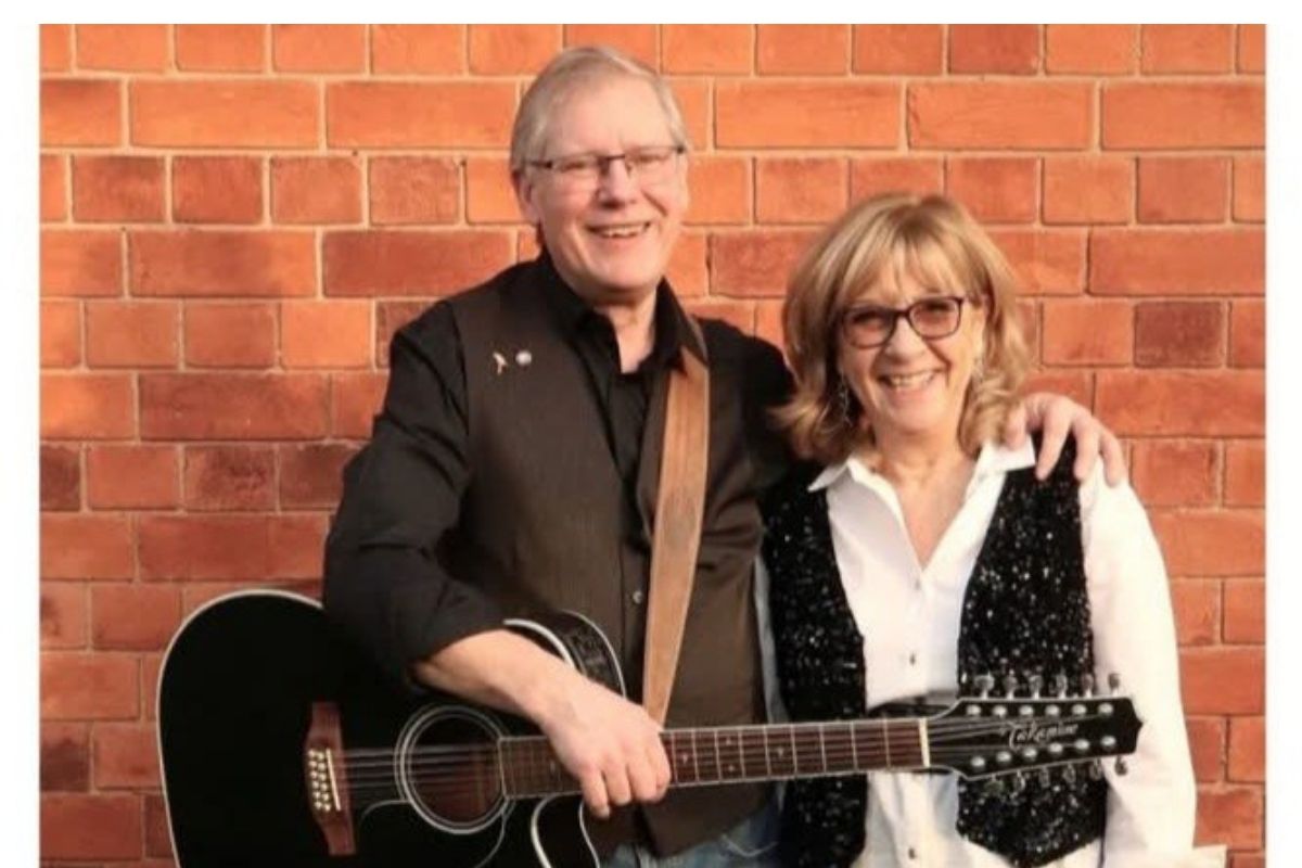 Two people standing side by side in front of a brick wall, with one person holding a black acoustic guitar