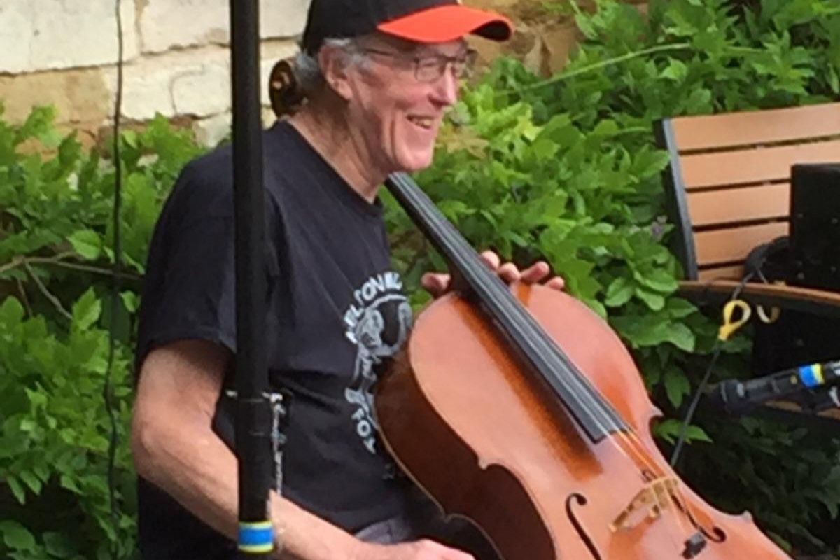 A person seated outdoors holding a cello, with music stands, plants, and equipment visible around them.