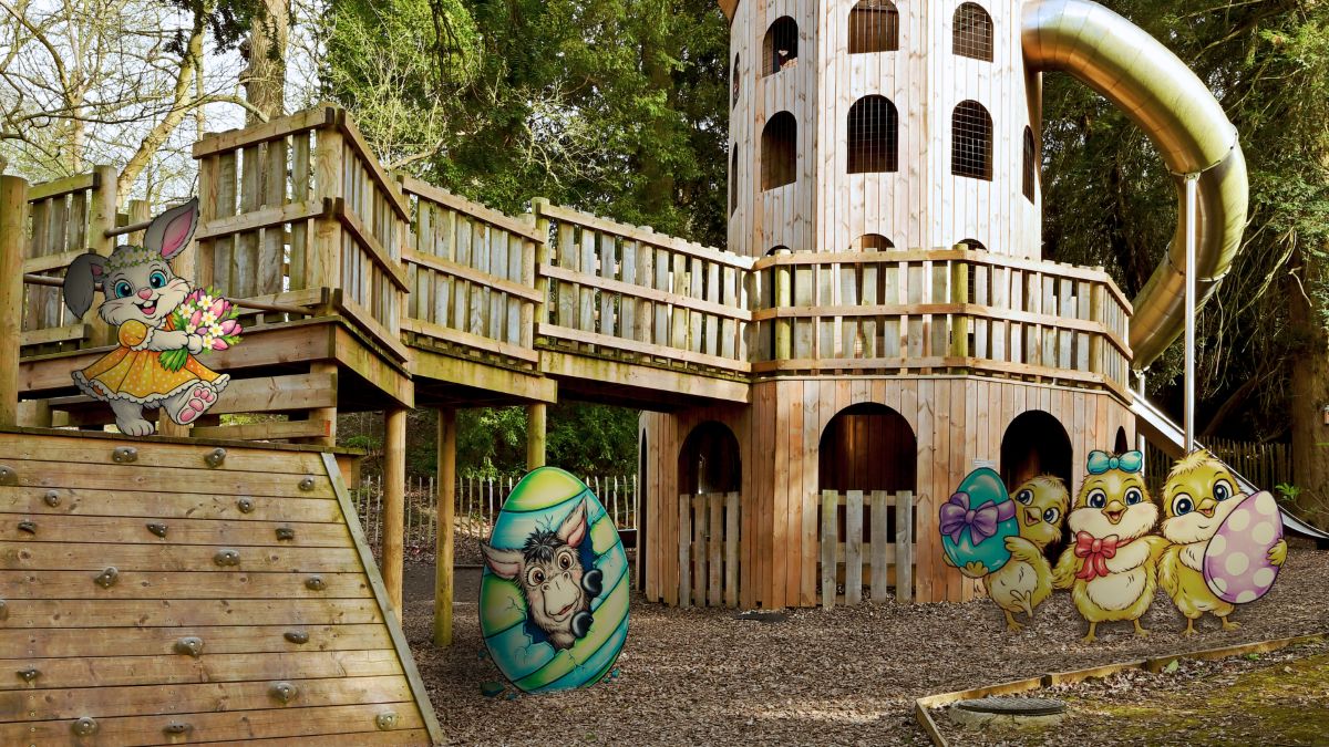 A wooden outdoor play structure with a tower, slide, and raised walkways in a wooded area. Cartoon Easter characters are added to the scene, including a bunny holding flowers on the left, a decorated egg with a cow illustration in the centre, and three chicks holding colourful Easter eggs on the right.