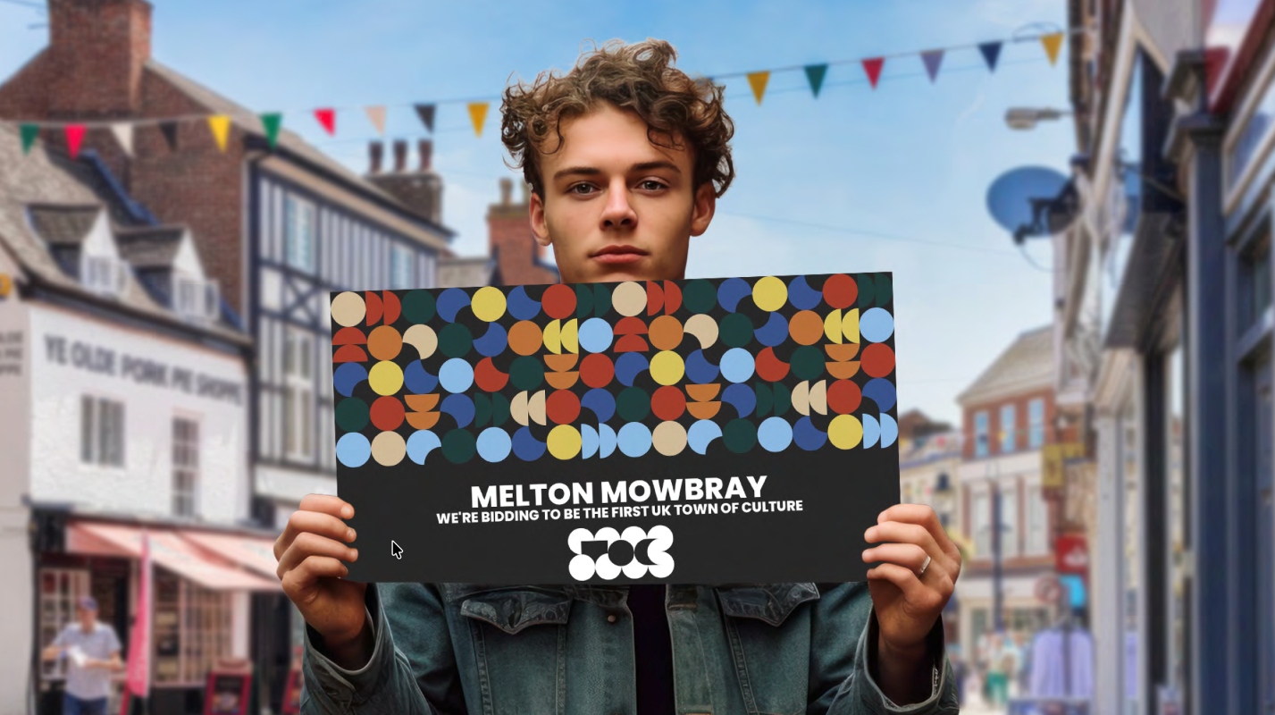 A young man stands in a colourful town street holding a large sign that reads “Melton Mowbray – We’re bidding to be the first UK Town of Culture.” The sign features a bold geometric pattern of multi coloured circles. Bunting hangs above the street and traditional shopfronts appear in the background