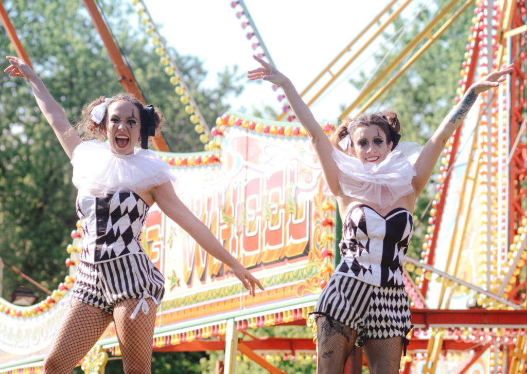 Two performers in black-and-white harlequin-style costumes with ruffled collars stand on an outdoor stage, arms raised. Behind them is a brightly decorated fairground ride with colorful lights and signage, framed by trees in daylight.