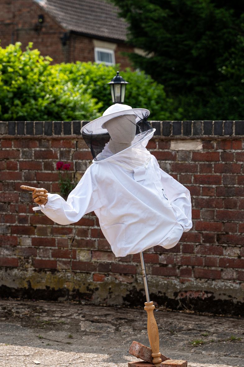 A mannequin dressed in a white beekeeping suit and veiled hat stands mounted on a pole, positioned in front of a red brick wall. The mannequin’s wooden hand points to the left. Behind the wall are green hedges, a lantern-style light post, and the upper part of a house with a tiled roof, all in soft outdoor daylight.