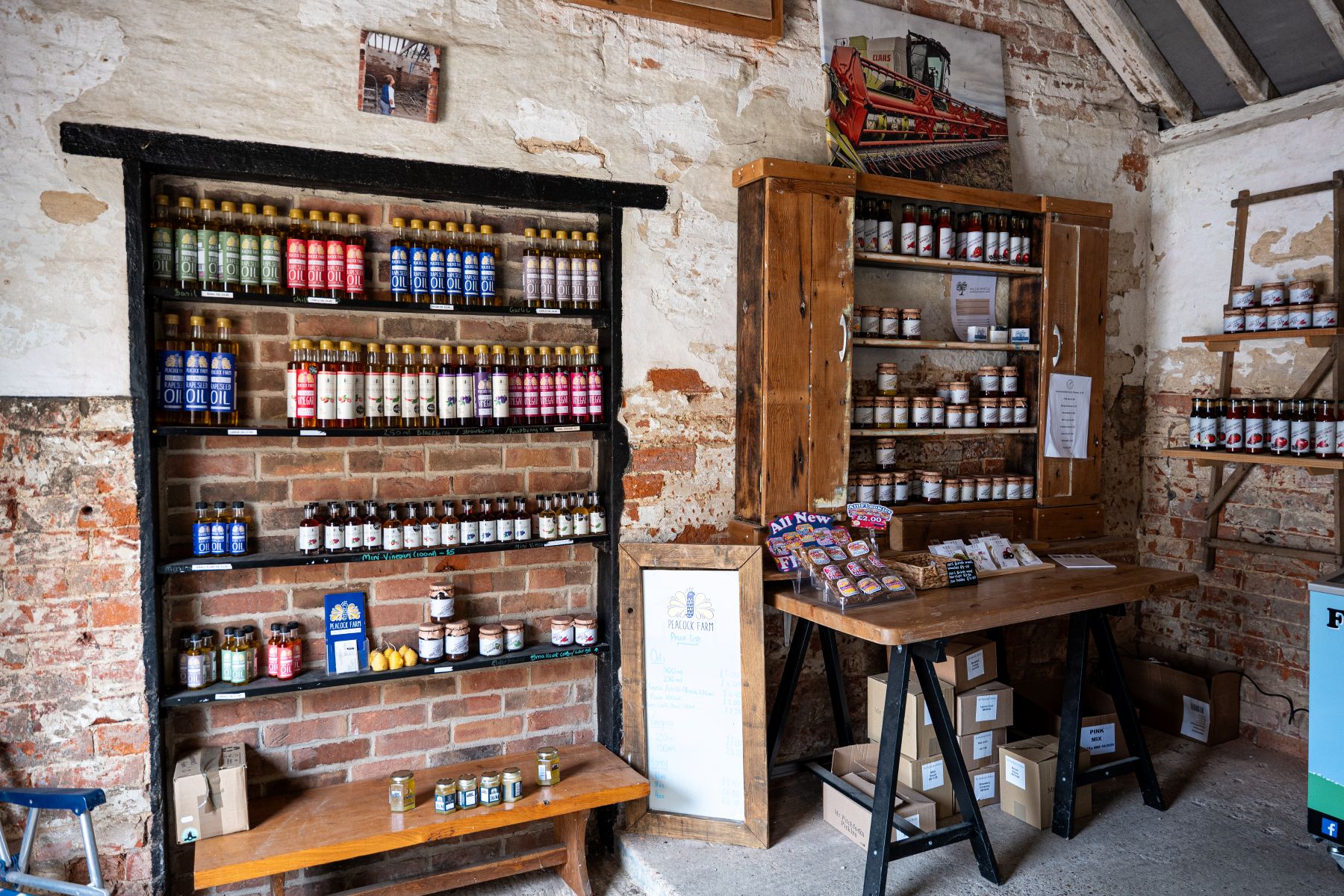 Interior of a rustic farm shop with exposed brick and plaster walls, displaying shelves of bottled oils, vinegars, and preserves. Wooden cabinets and tables hold jars, sauces, and packaged goods, with a handwritten price list board and stacked boxes beneath. A photo of farm machinery hangs above the shelving, adding to the agricultural setting.