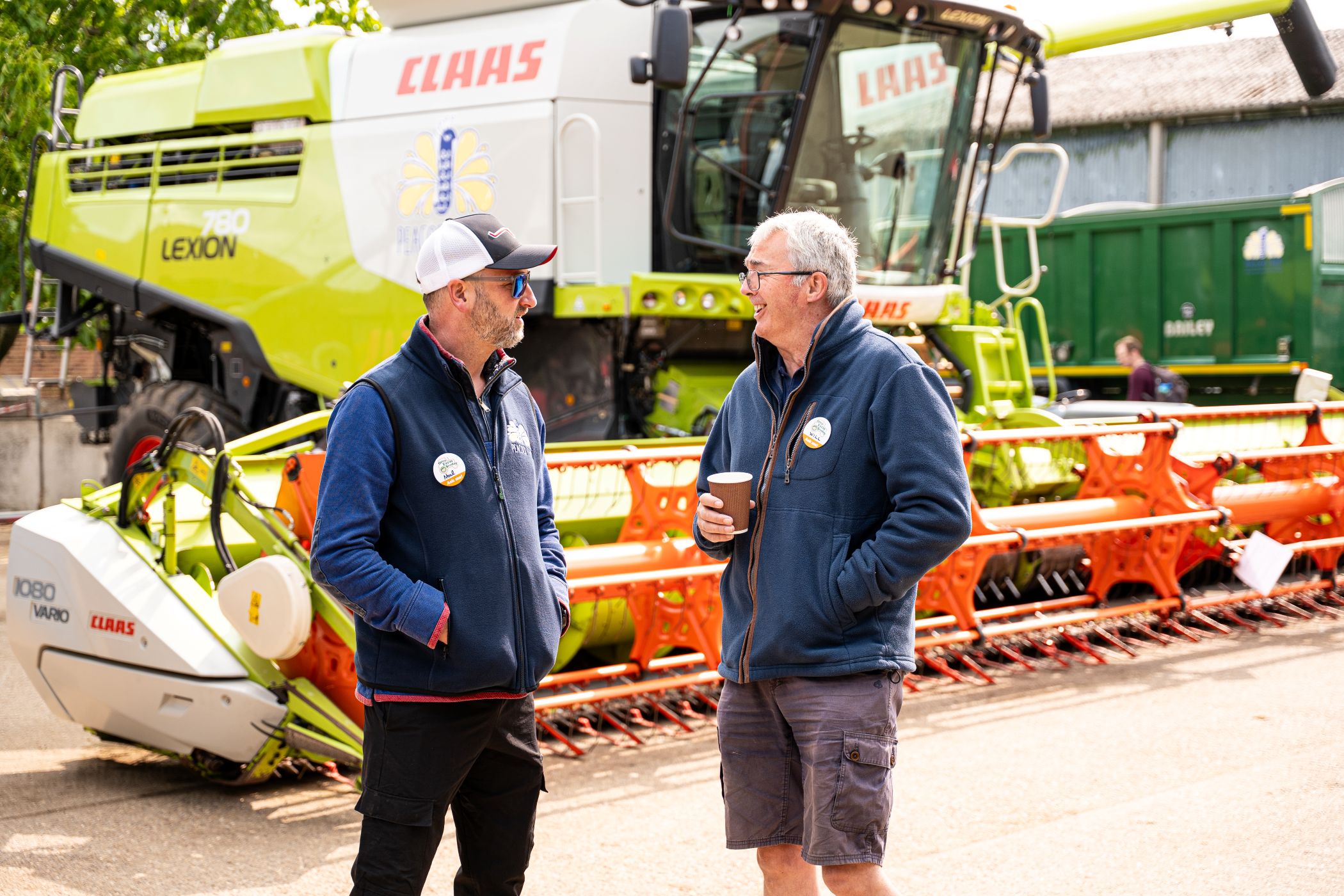 Two people stand talking in a farmyard in front of a large green and white CLAAS LEXION 780 combine harvester. Both wear casual outdoor clothing with badges, and one holds a takeaway coffee cup. The bright orange cutting header is visible in the foreground, with farm buildings and machinery in the background under sunny daylight.