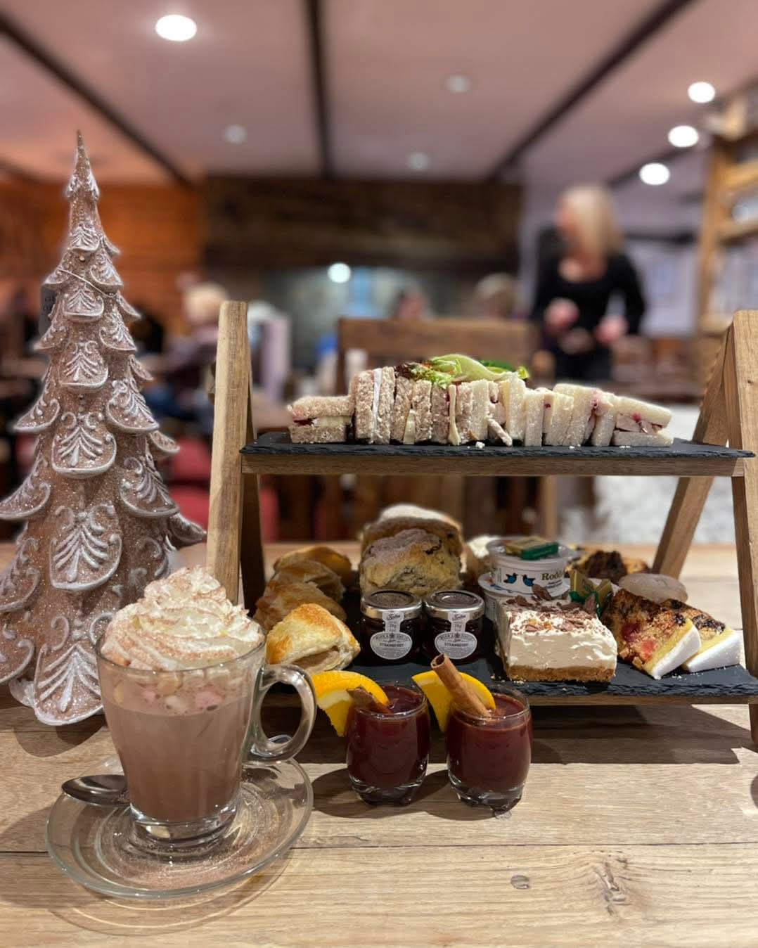 Festive afternoon tea with sandwiches, cakes, hot chocolate and Christmas treats served beside a decorative Christmas tree.