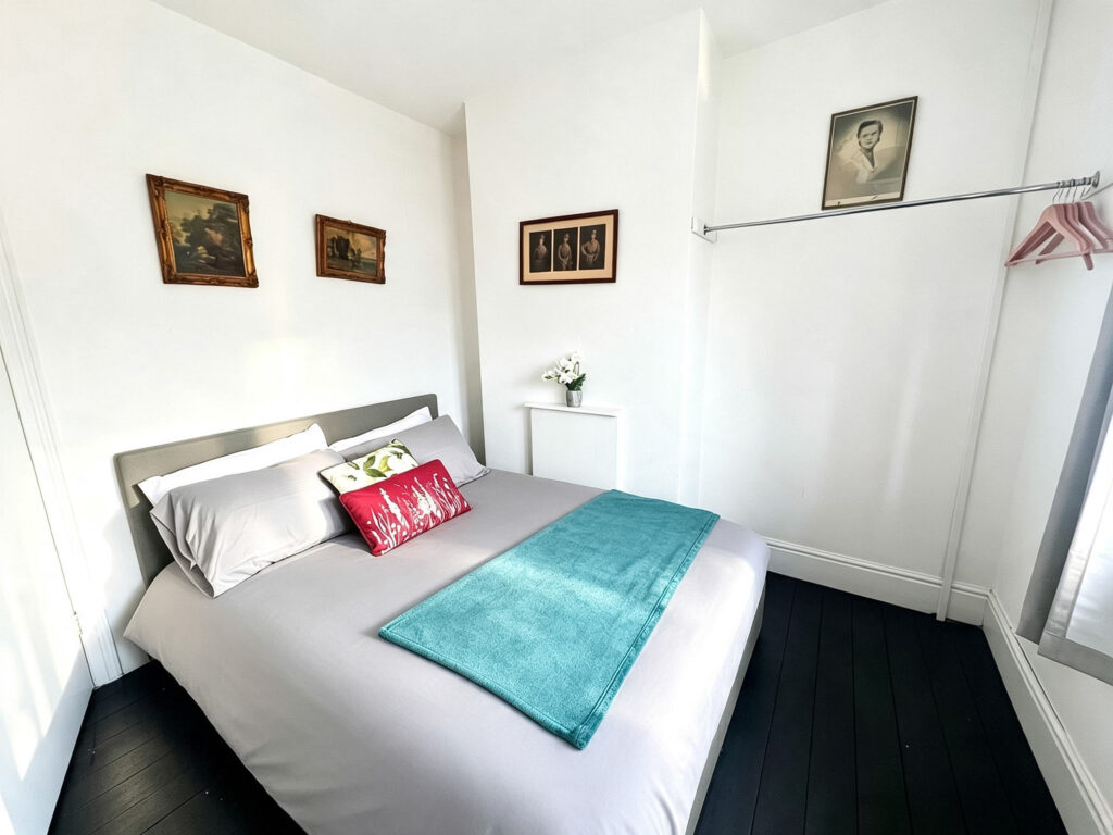 Bright double bedroom with grey bedding, turquoise throw, white walls, and framed artwork.