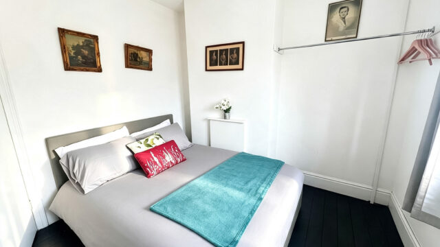 Bright double bedroom with grey bedding, turquoise throw, white walls, and framed artwork.