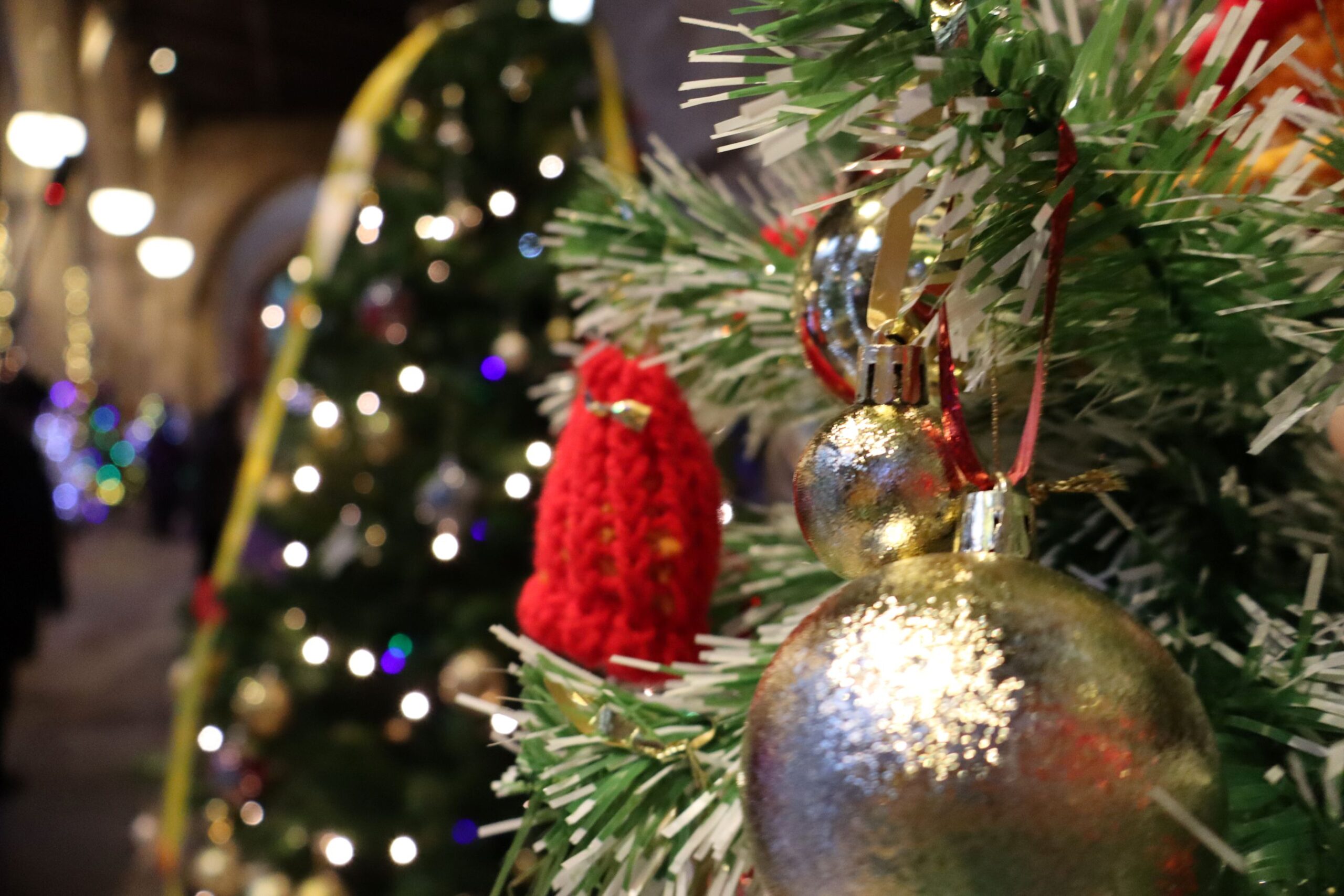 Close-up of silver and gold baubles, red knitted decorations and white-tipped branches on an illuminated Christmas tree, with softly blurred festive lights and trees in the background.